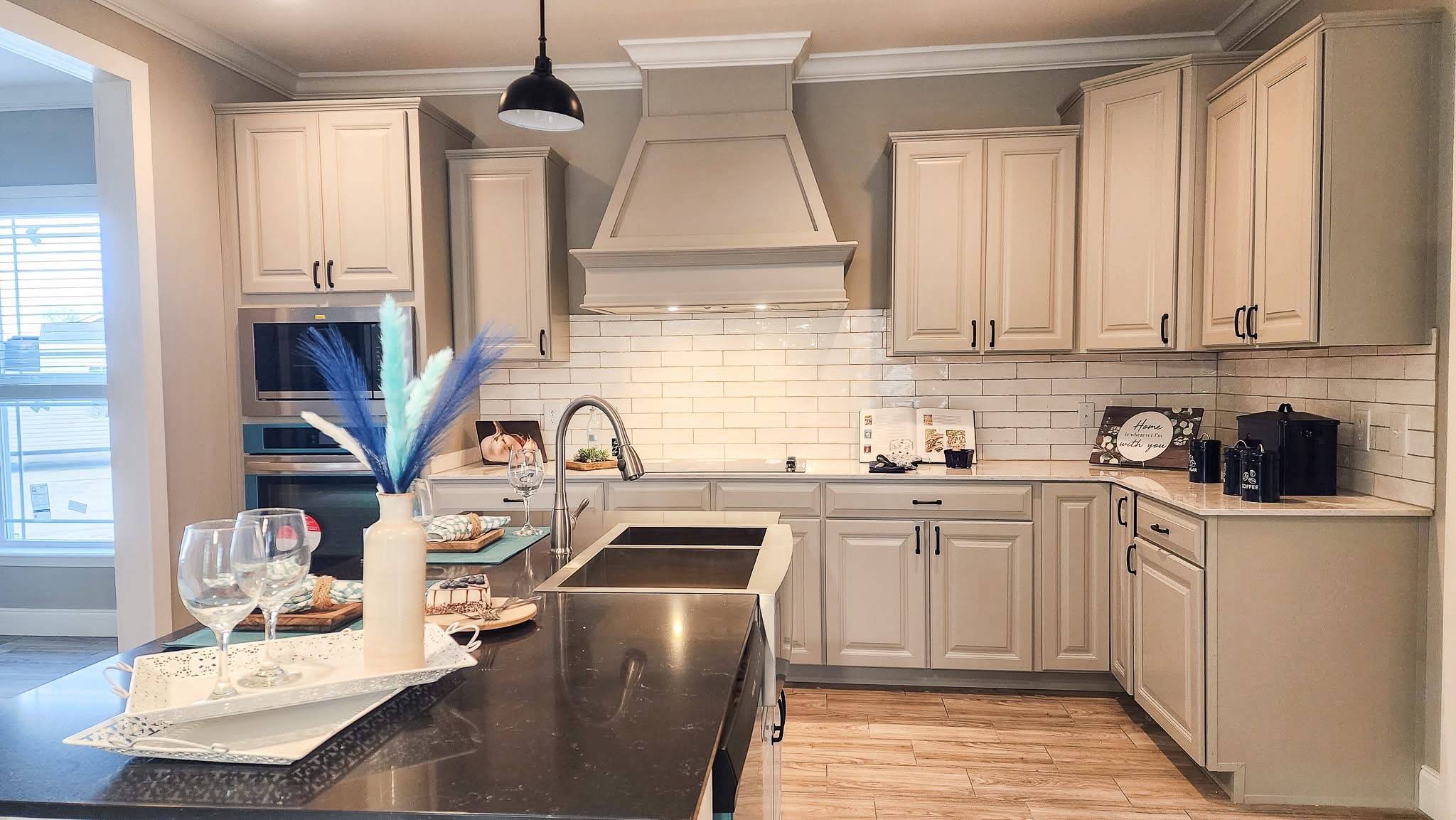 A modern kitchen with cream cabinets, subway tile backsplash, and a black countertop. A vase with decorative feathers and wine glasses are on the counter.