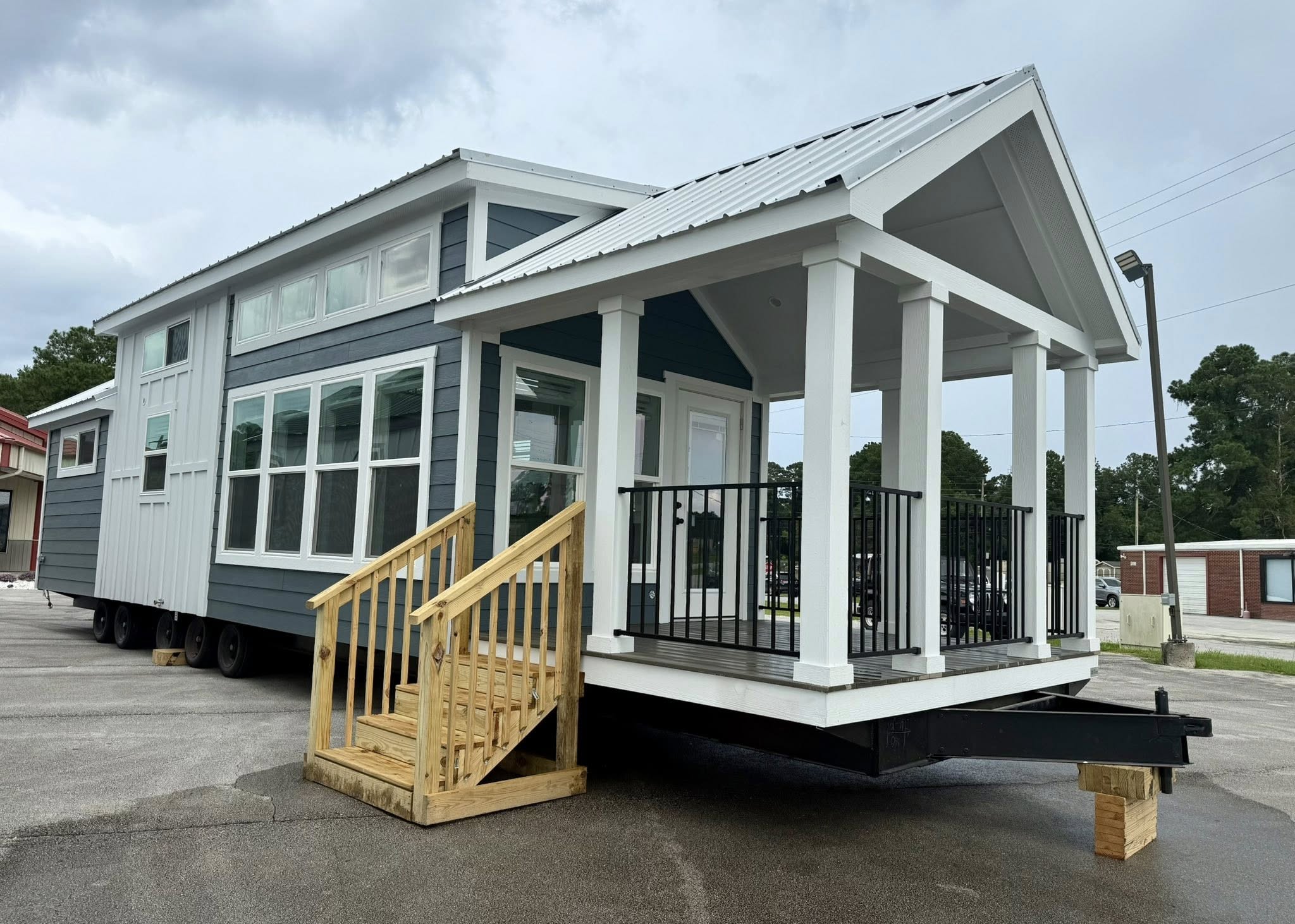 A modern tiny house with blue and white siding, large windows, and a small porch. Wooden steps lead to the entry. Overcast sky, residential area.