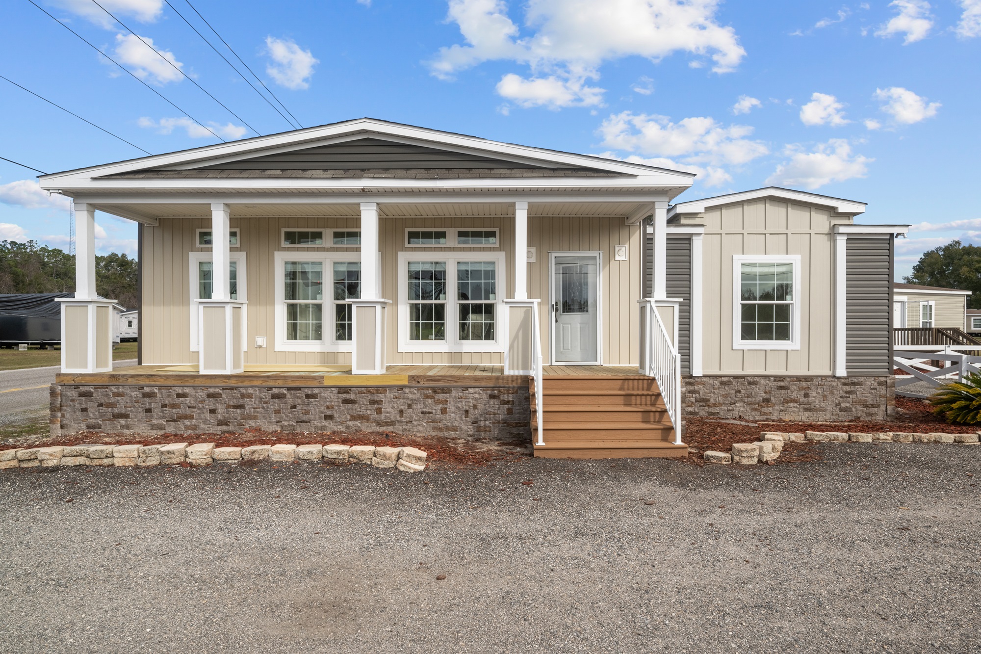 Single-story manufactured home with beige siding, white trim, and stone base. Features a front porch with steps, large windows, and a blue sky.