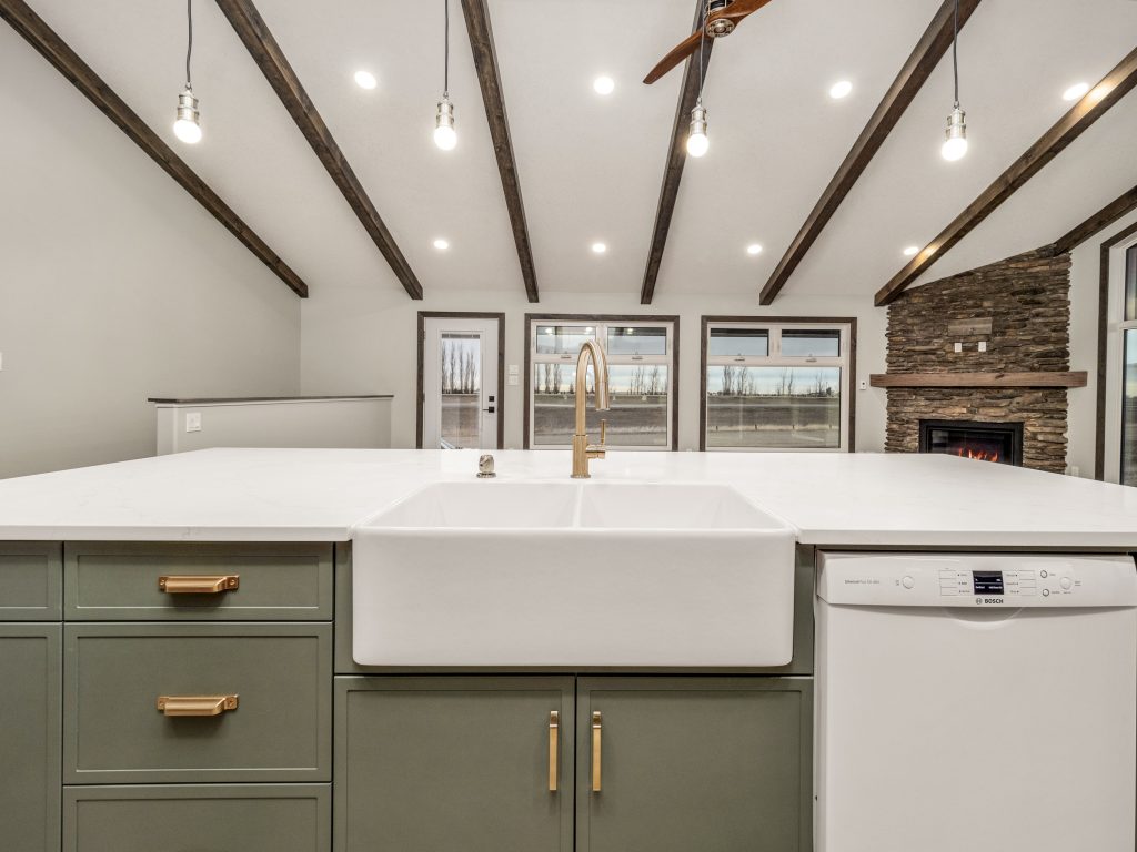Spacious kitchen with green cabinets, a white farmhouse sink, and gold fixtures. Exposed beams and stone fireplace create a rustic yet modern feel.