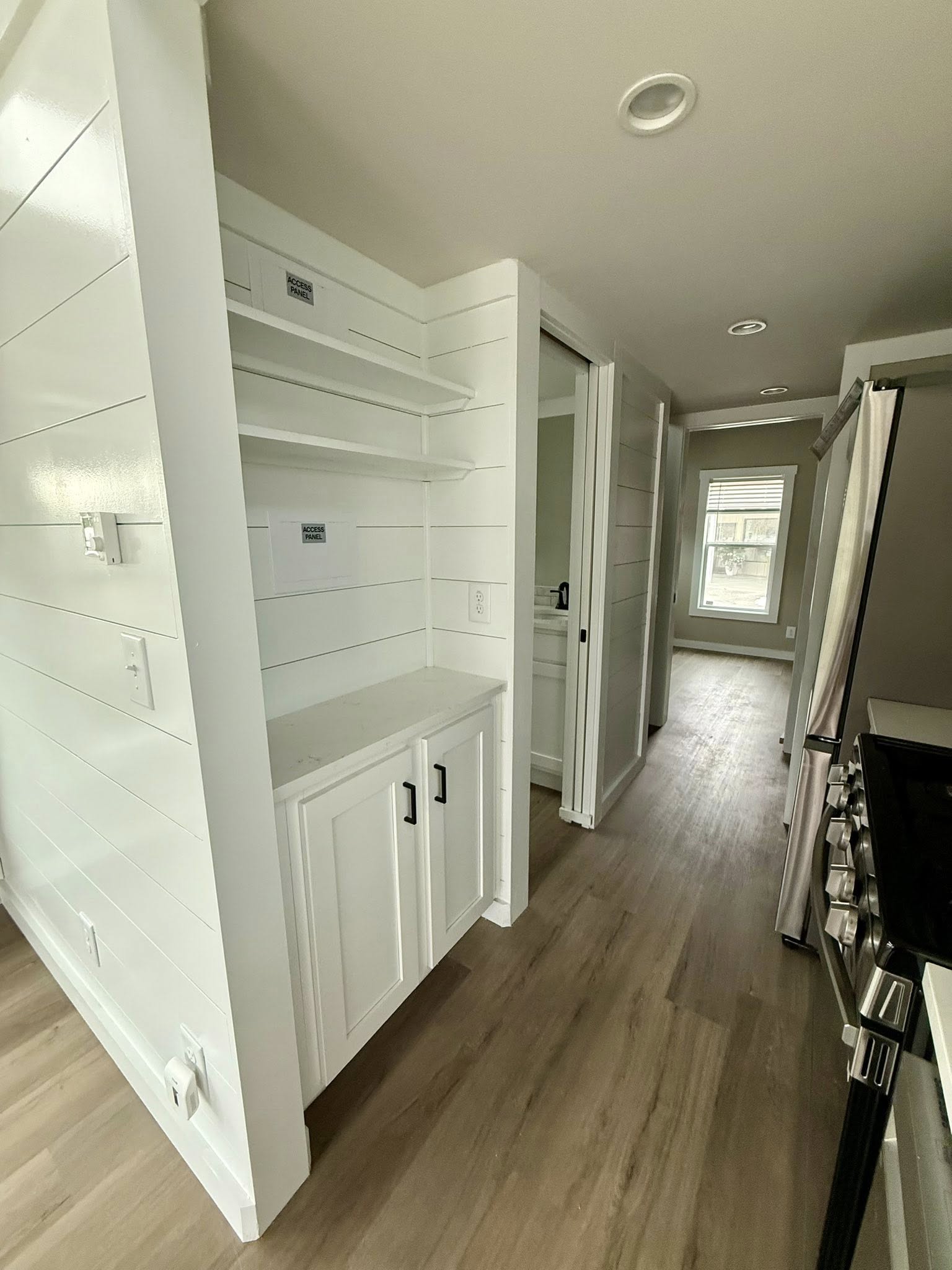 A narrow, modern hallway with white shiplap walls and light wood flooring. It features open shelves and cabinets, leading towards a sunlit window.