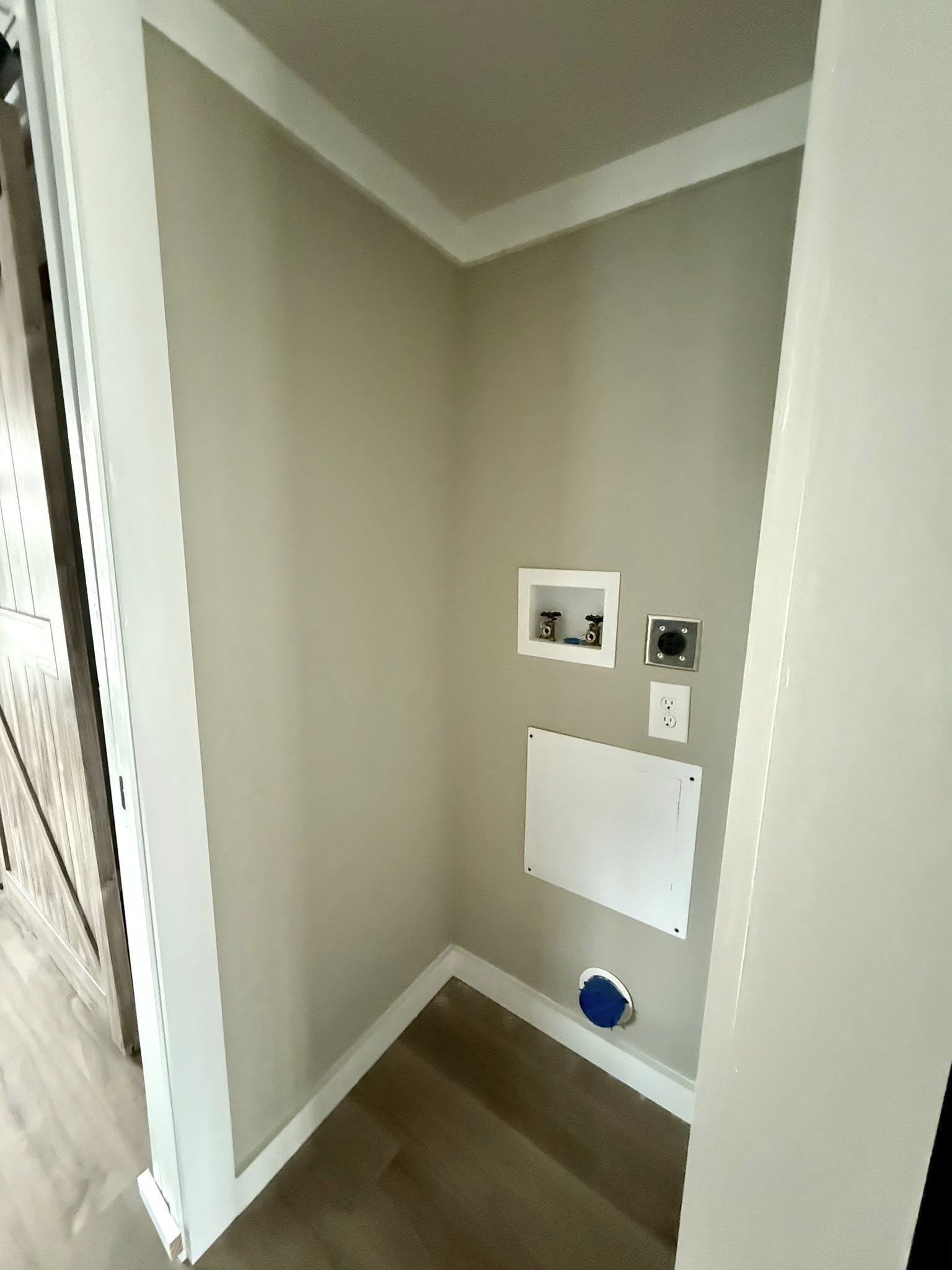 A small laundry nook with water hookups and an electrical outlet on a gray wall. The wooden floor adds warmth to the clean, minimalistic space.