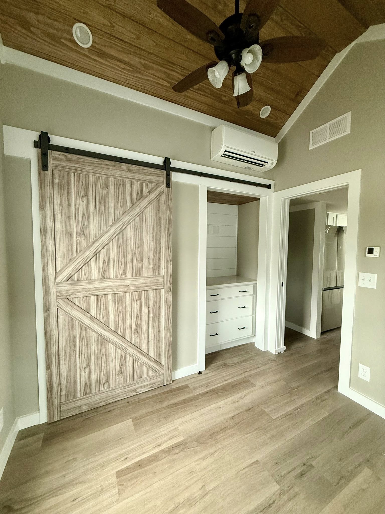 Cozy room with a rustic sliding barn door, light wood flooring, and vaulted ceiling with fan. Built-in drawers and AC unit above the door add functionality.