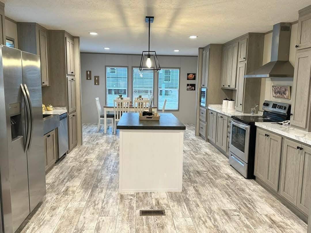 A spacious kitchen with gray wood cabinets, a central island, stainless steel appliances, and a dining area with white chairs under soft lighting. Modern and inviting.
