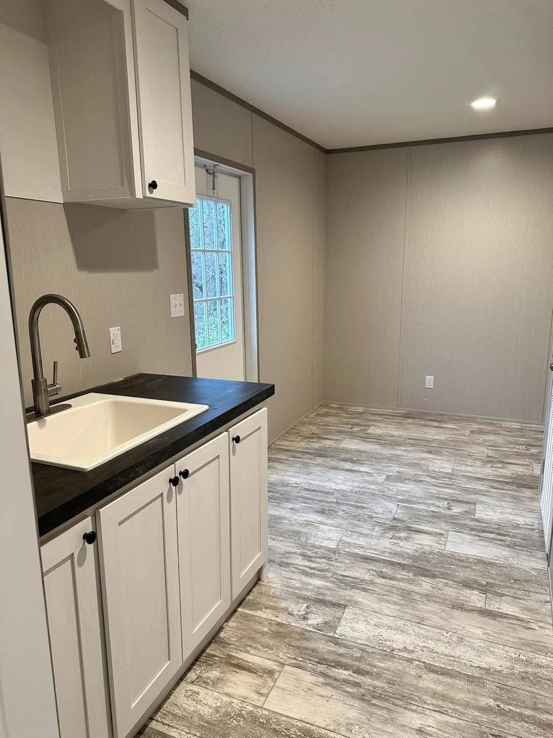 Compact kitchen space with white cabinets and dark countertop, featuring a sink and faucet. The room has gray wood-look flooring and a soft, neutral tone.