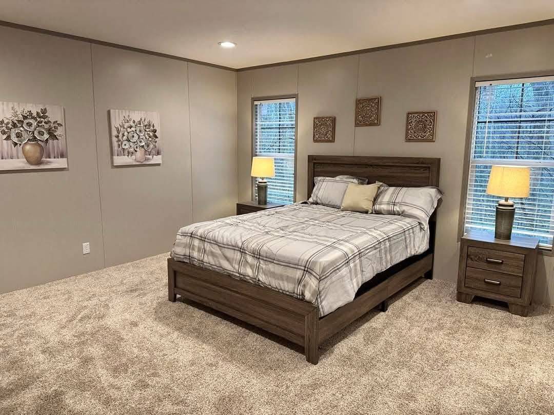 A cozy bedroom with a plaid-sheeted bed centered between two windows, each with a lit lamp. Neutral walls feature floral artwork, creating a serene vibe.
