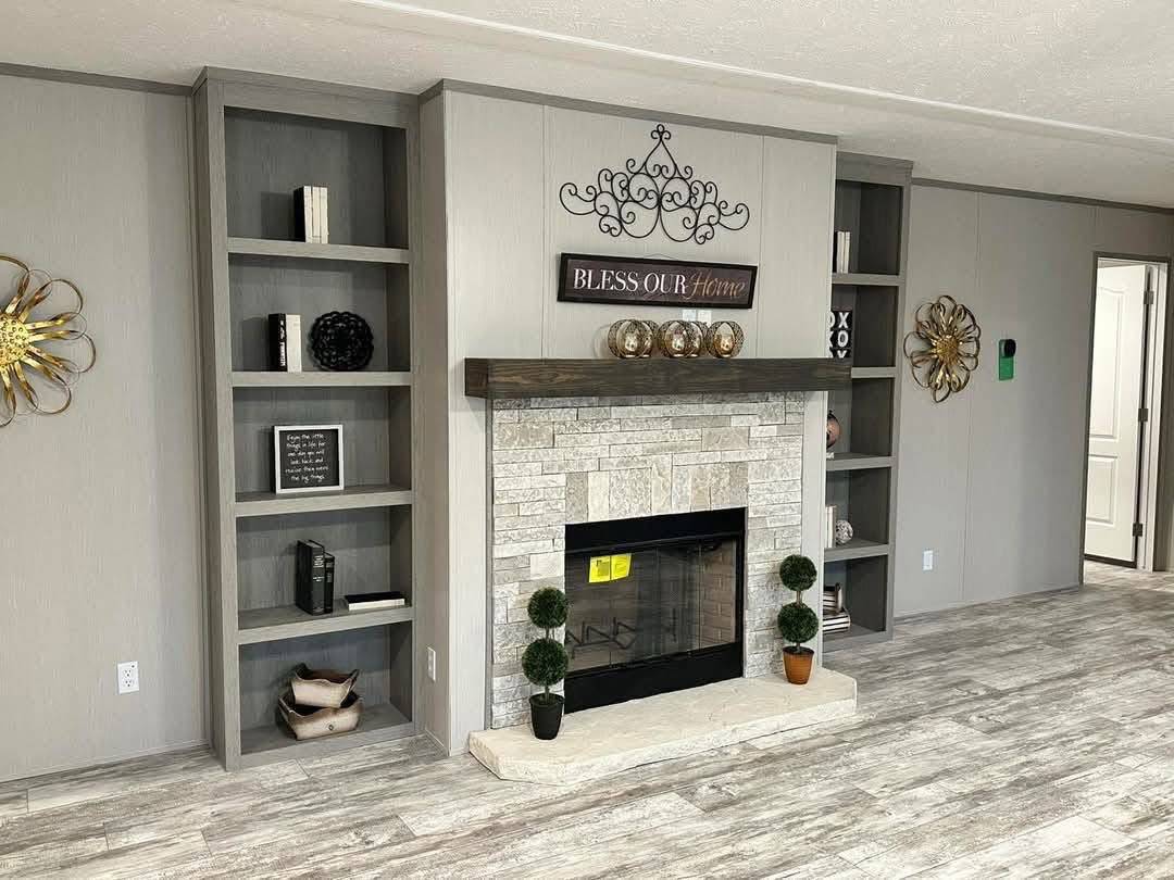 Modern living room with stone fireplace, decorative shelving, and wall art saying "Bless Our Home." Neutral tones and cozy ambiance.