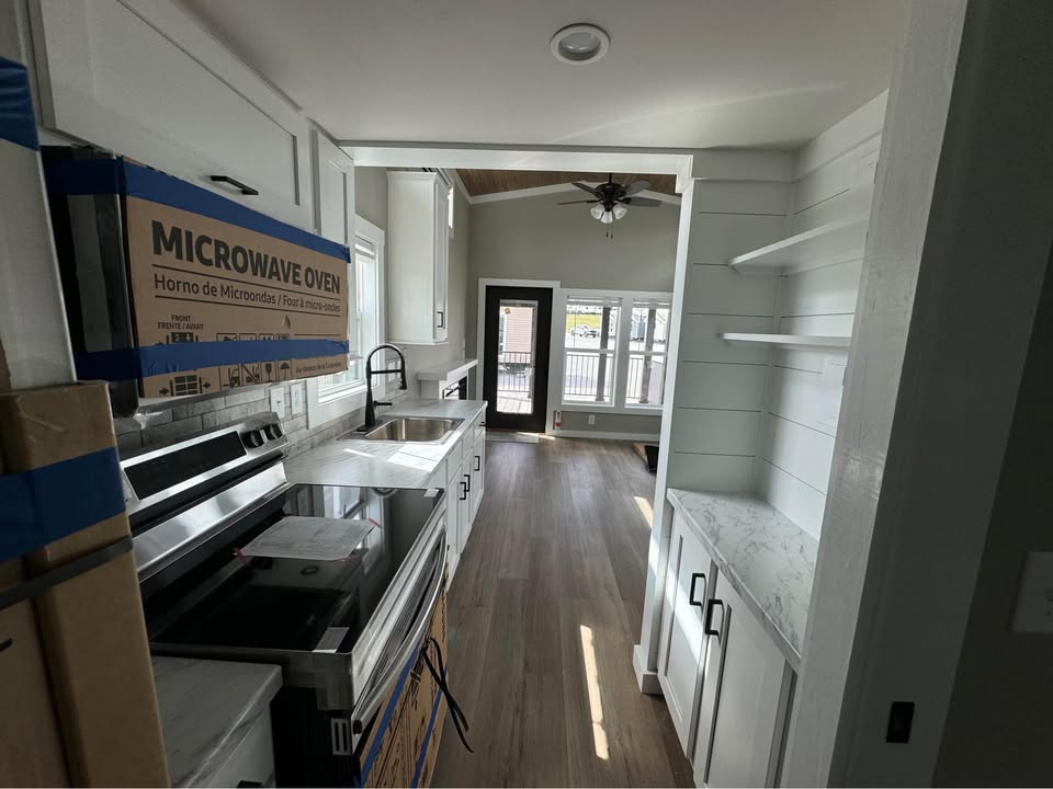 A narrow, modern kitchen with white cabinets, a microwave, and a sink on the left. Light wood flooring leads to a bright room with large windows.