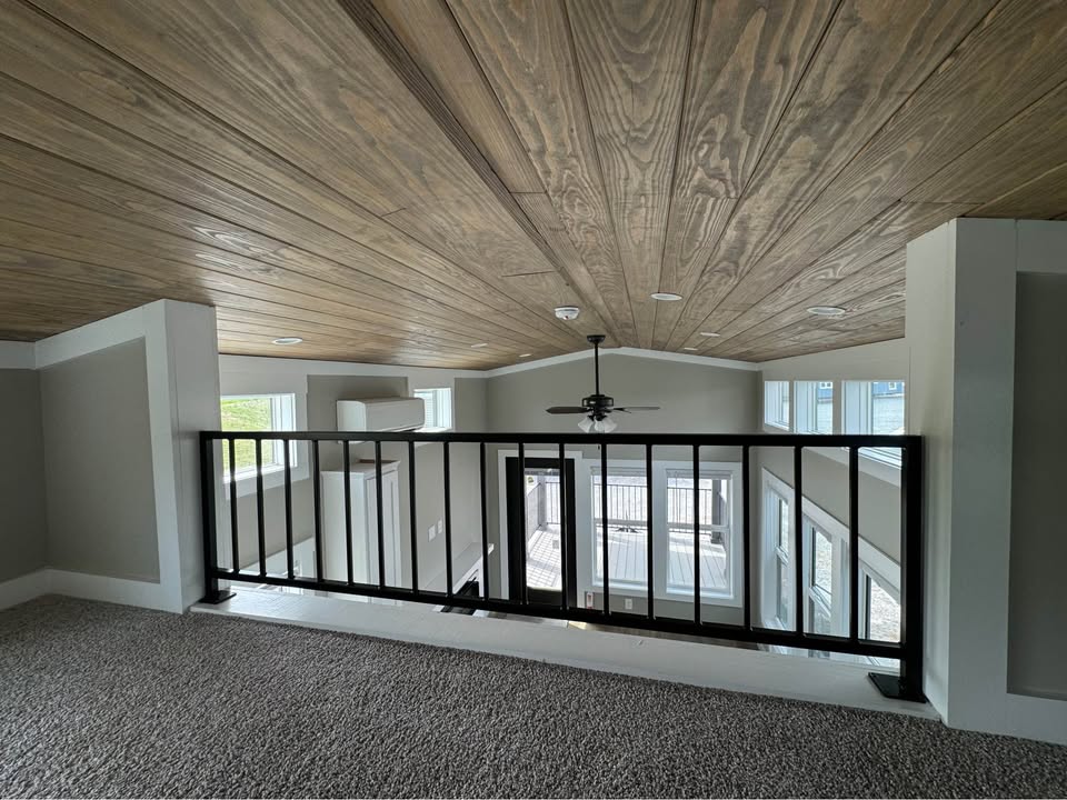 Loft interior with wooden ceiling, black railing, and gray carpet. Ceiling fan visible above, windows below provide natural light, creating an airy feel.