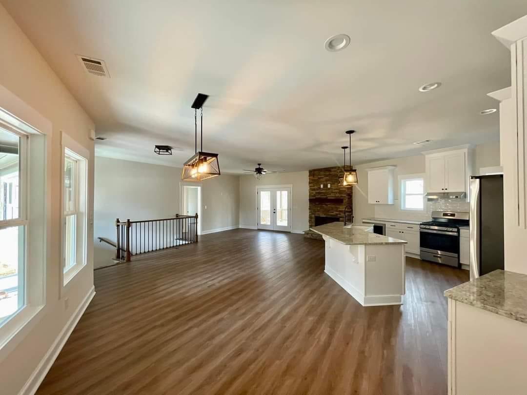 Spacious open-concept kitchen and living area with wooden floors, white cabinets, and stone fireplace. Large windows and modern lights create a warm ambiance.