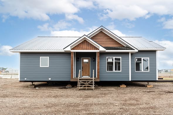 Single-story prefabricated home with blue siding, brown accents, and a metal roof, set on a dirt lot. The sky is partly cloudy, suggesting a calm day.