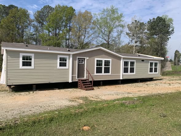 A beige mobile home with white trim and a small red staircase sits on a gravel path. It's surrounded by a grassy area and trees under a blue sky.