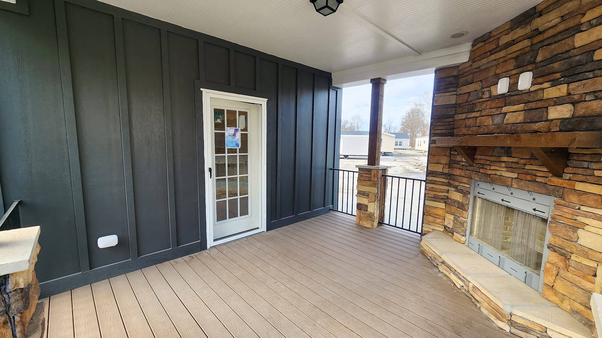Covered porch with a light wood floor and dark paneled walls, featuring a stone fireplace with a chimney. A white glass door leads inside. Cozy and inviting.
