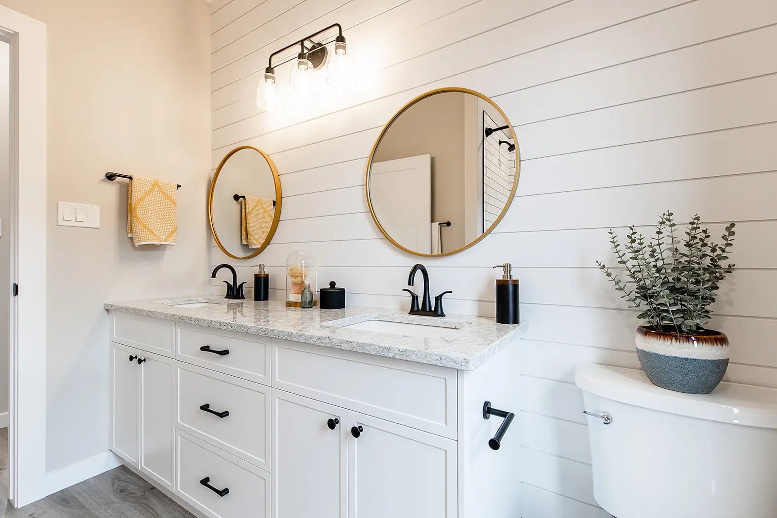 Modern bathroom with dual sinks, white cabinets, and round mirrors on a shiplap wall. Bright lighting, black faucets, and a potted plant add elegance.