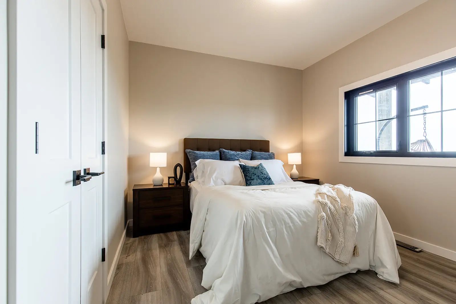 A cozy bedroom with beige walls, featuring a neatly made bed with white linens and blue pillows. Two bedside tables with lamps add warmth. A large window lets in natural light.