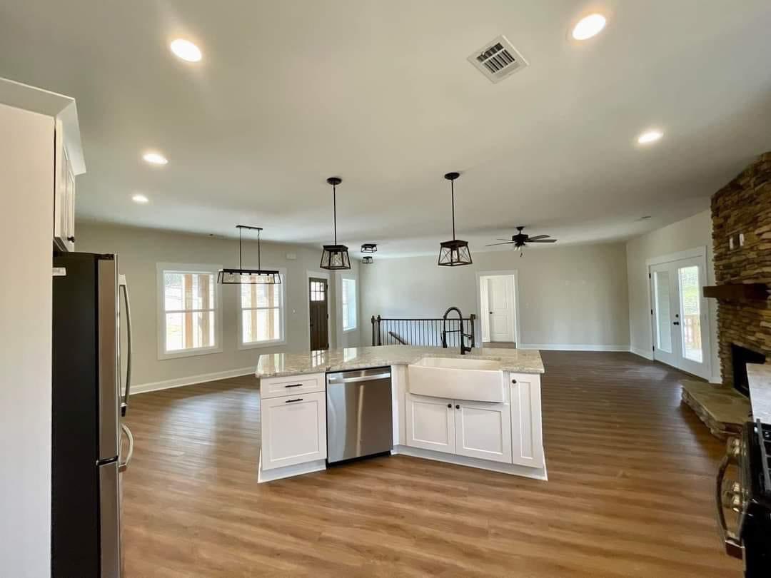 Spacious open-concept kitchen and living area with wooden floors, centered island, modern lighting, stone fireplace, and large windows, conveying warmth.