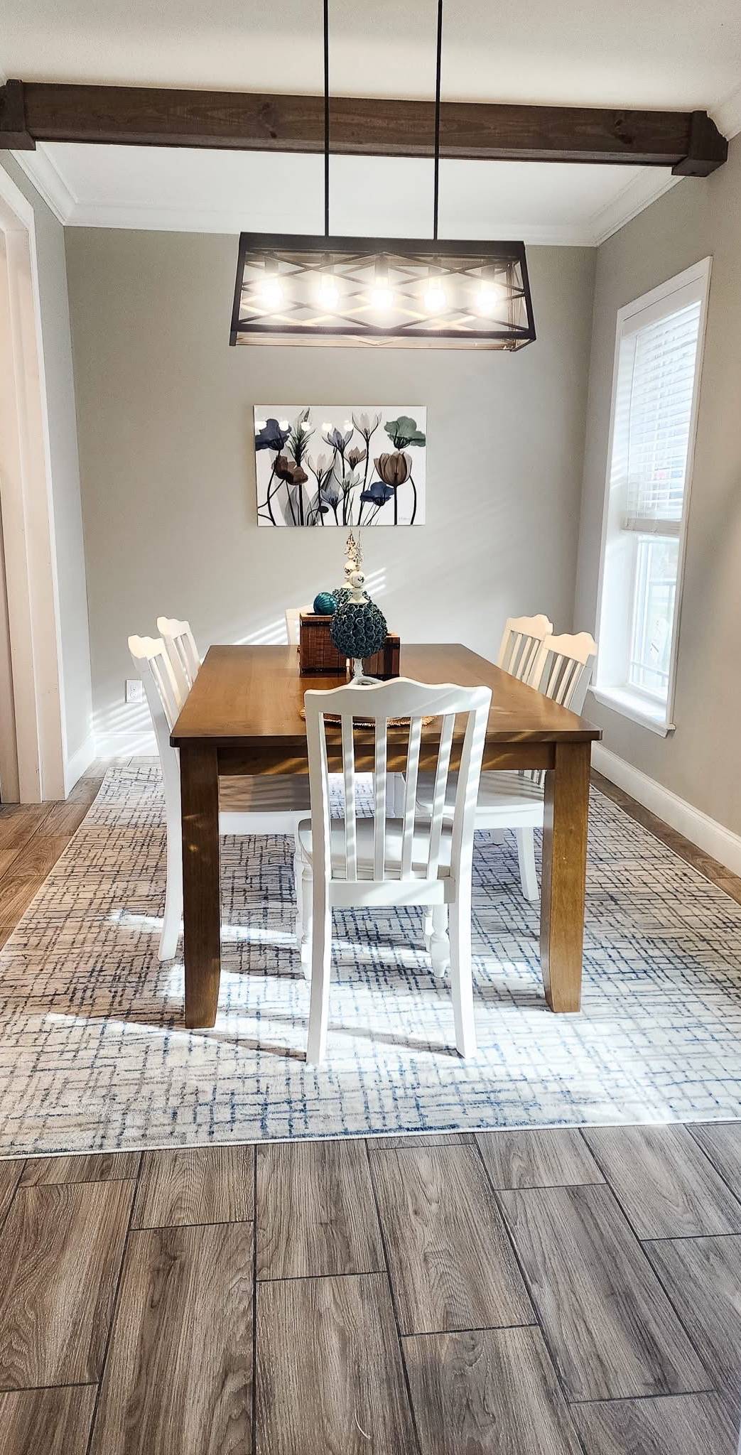 A cozy dining room with a wooden table and four white chairs on a patterned rug. A modern light fixture hangs above, and floral artwork decorates the wall.