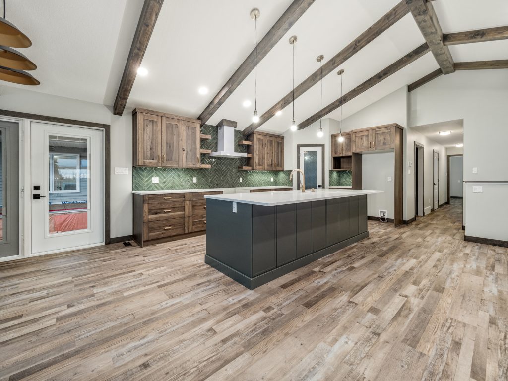Spacious kitchen with vaulted ceiling and exposed beams. Features a central island, wooden cabinets, green tile backsplash, and wood flooring.