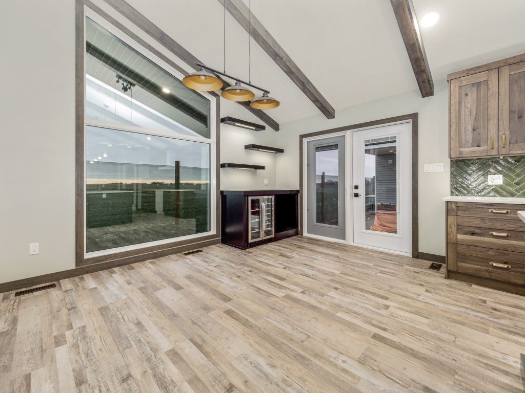Spacious room with large windows, wood beam ceiling, and light wood flooring. Features include a small bar, floating shelves, and a modern pendant light.