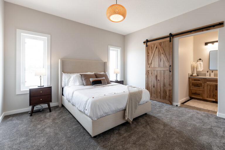 A cozy bedroom features a beige bed with white linens, flanked by two wooden nightstands with lamps. A rustic sliding barn door opens to a bathroom.