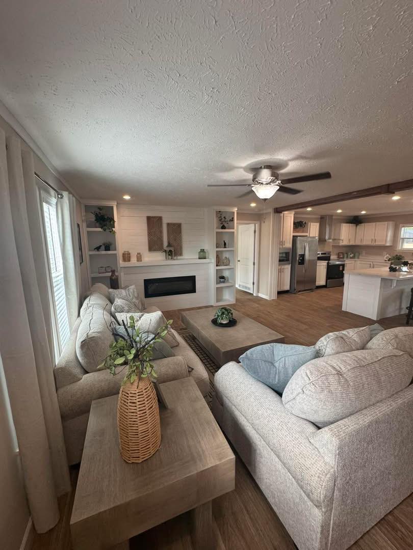 Cozy living room with beige sofas, a wicker vase, and a large coffee table. A ceiling fan and a fireplace enhance the inviting, serene ambiance.
