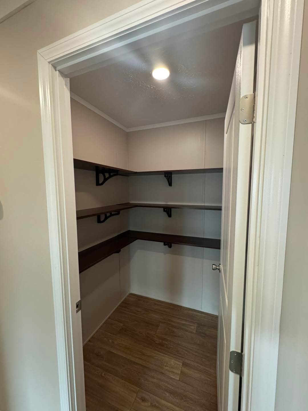 A small walk-in closet with wooden shelves and light wood flooring. The shelves create an L-shape, maximizing storage. A soft ceiling light illuminates the space.