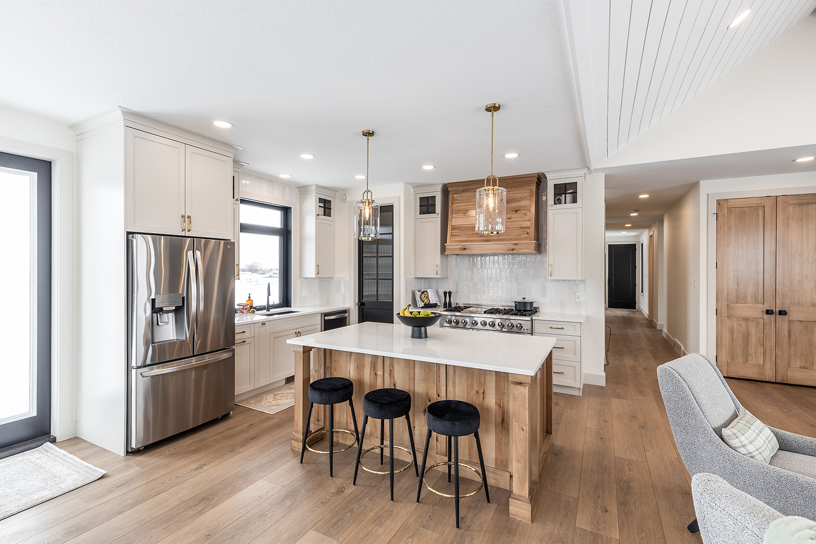 Modern kitchen with light wood floors, a central island with three black stools, stainless steel appliances, and pendant lights, exuding a cozy ambiance.