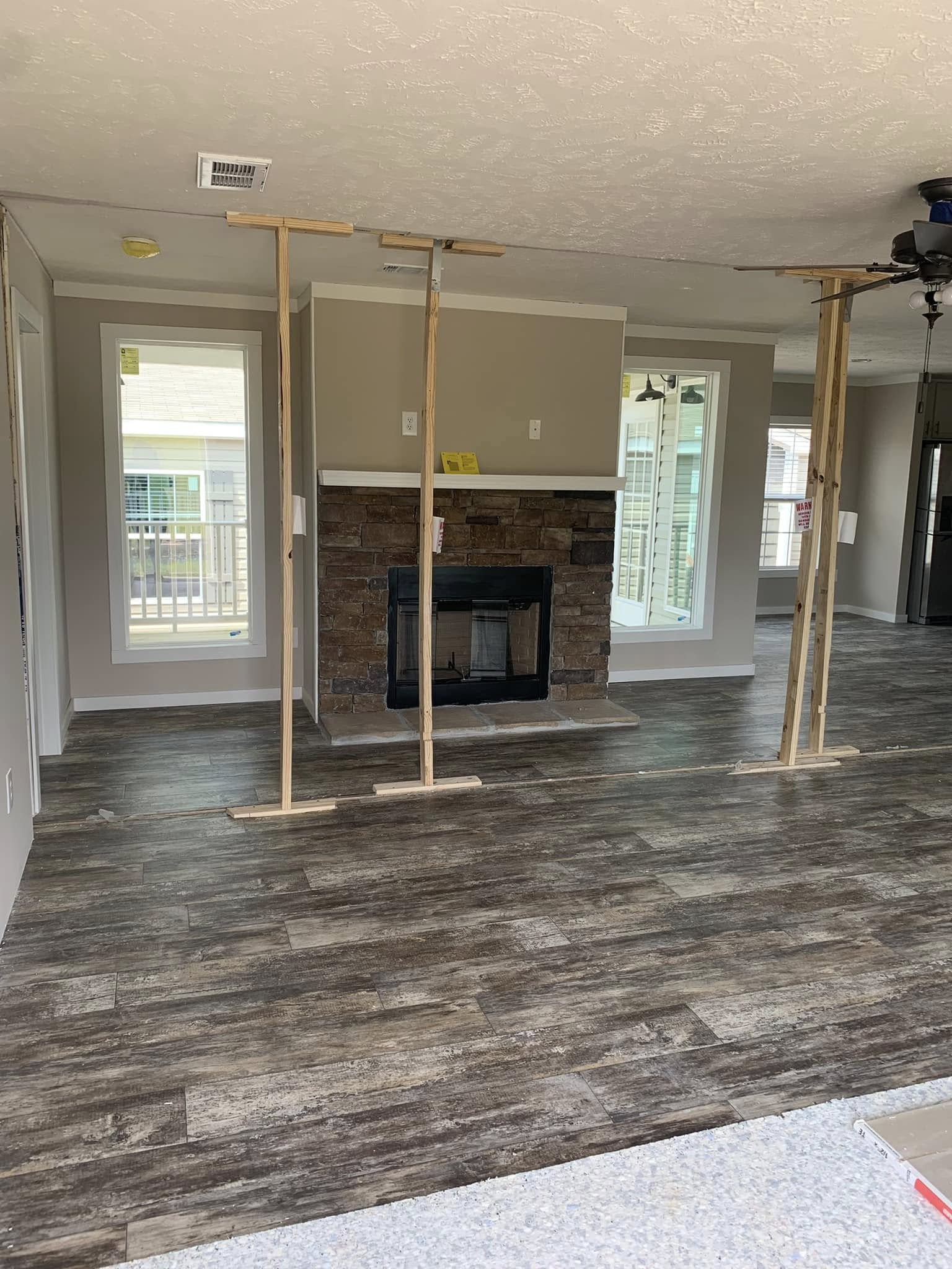 Spacious living room with dark wood flooring and a stone fireplace. Temporary wooden supports prop the ceiling. Bright windows frame a calm, unfinished view.