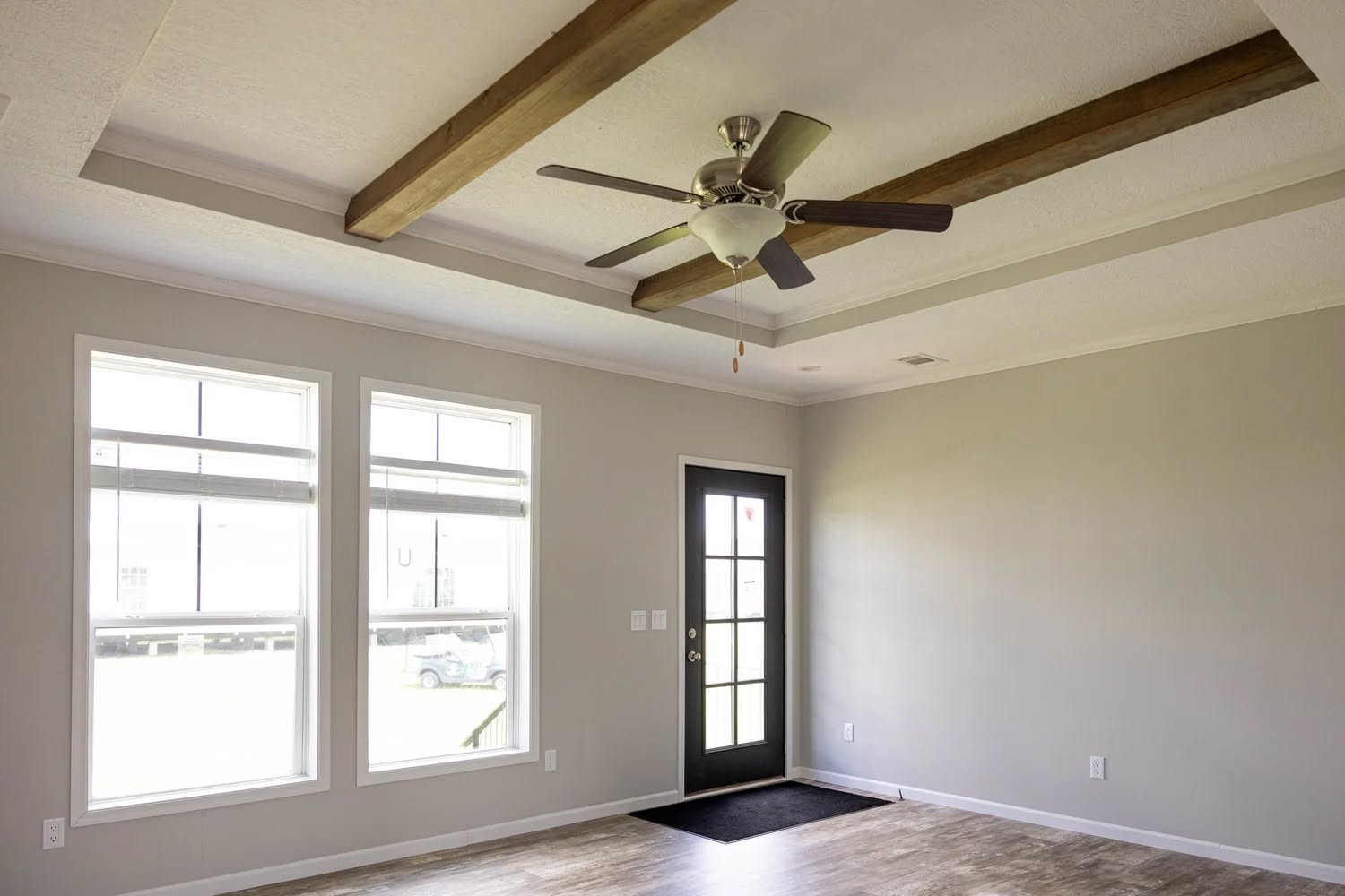 A bright, empty room with light gray walls, wooden ceiling beams, a ceiling fan, large windows, and a black door. The room has a modern, airy feel.