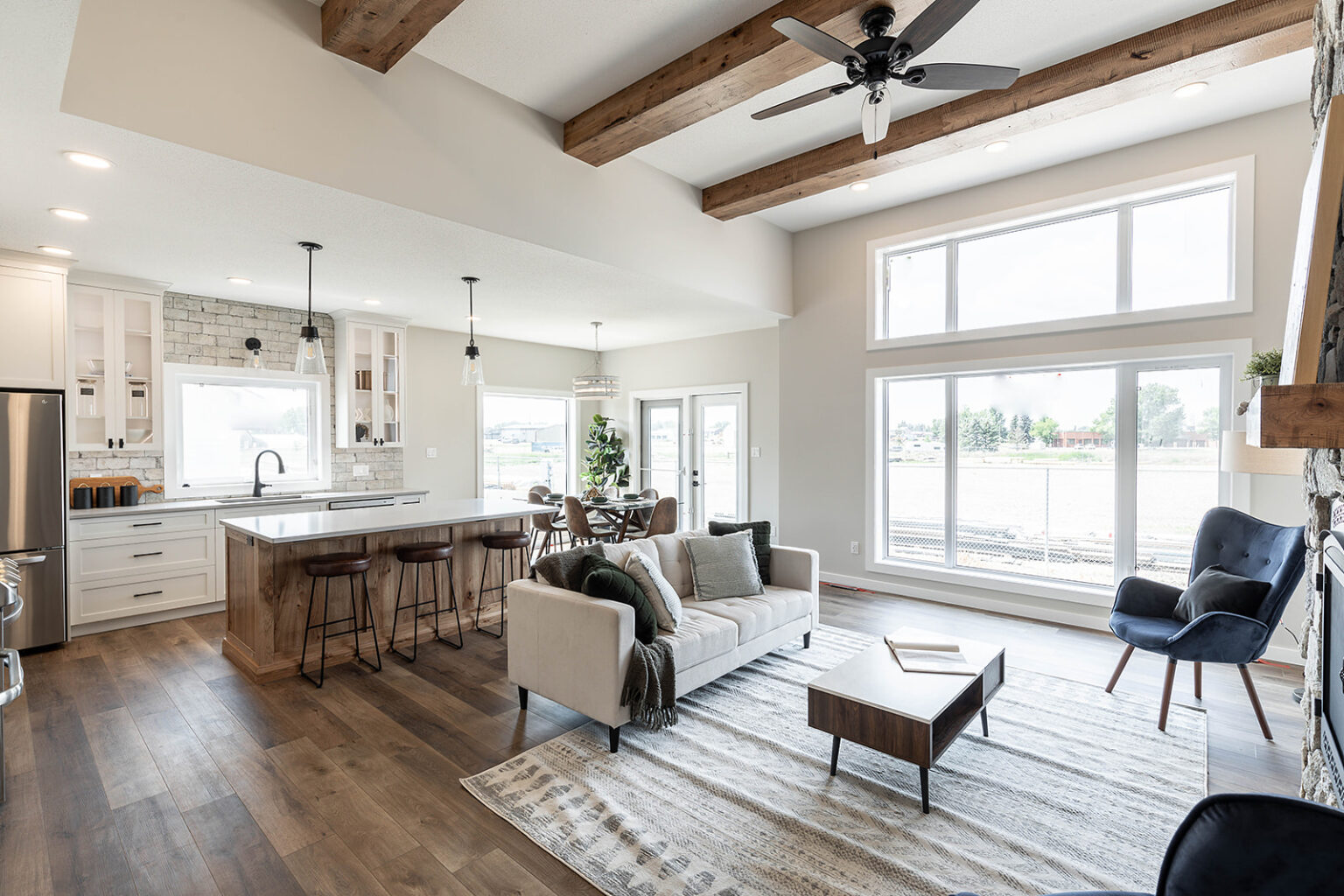 Spacious open-concept living room and kitchen featuring wooden beams, a ceiling fan, large windows, a cozy couch, dining table, and modern decor.
