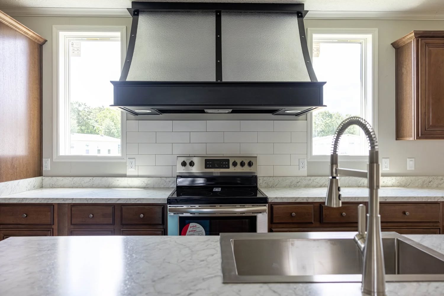 Modern kitchen with marble countertops, a stainless steel sink, and faucet in the foreground. A stove and large range hood are centered, flanked by windows.