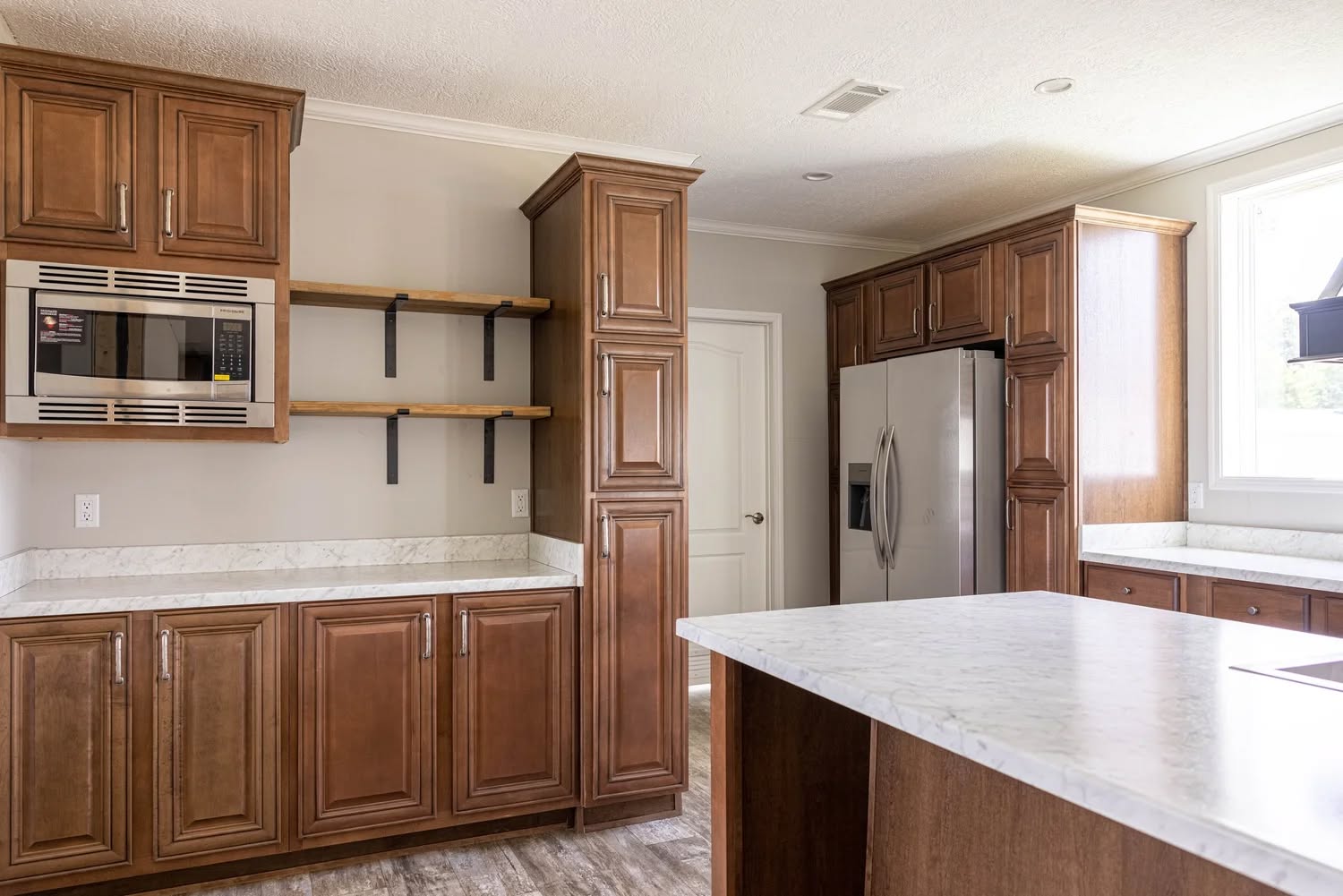 Modern kitchen with wooden cabinets, a built-in microwave, open shelves, and a stainless steel fridge. Bright marble countertops and a large island.