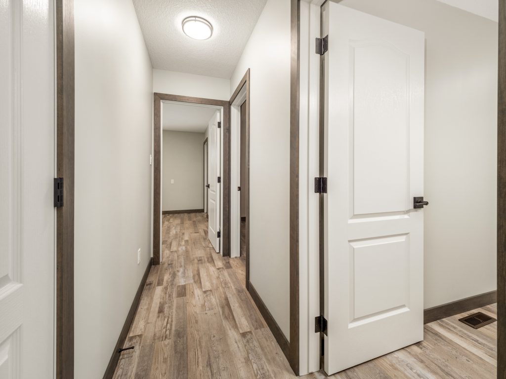 Wood-floored hallway with cream walls and dark trim, featuring multiple open doors leading to adjacent rooms. Ceiling light enhances the bright, clean atmosphere.