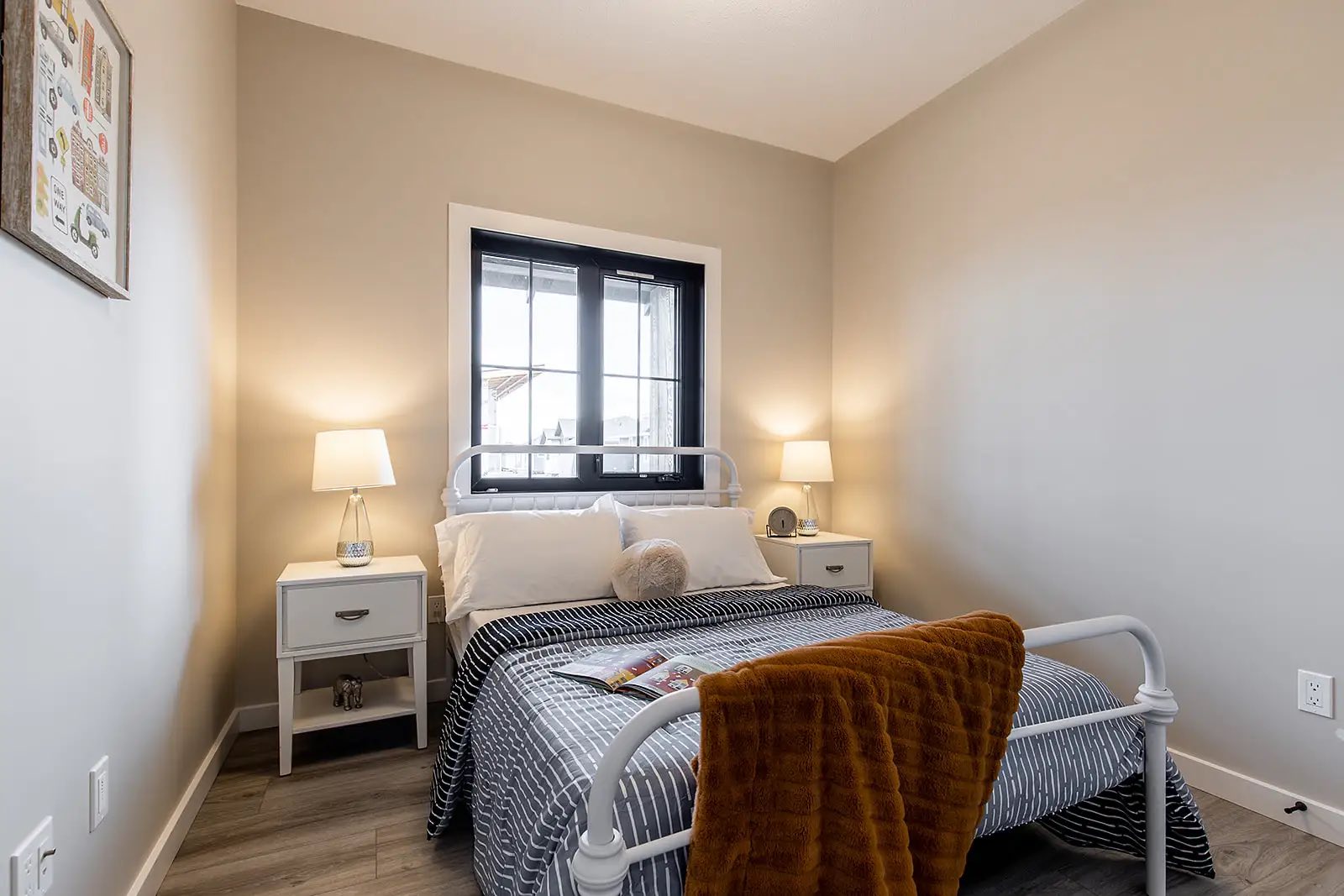 Cozy bedroom with a neatly made bed, gray striped bedding. Two side tables with lamps, a black-framed window, and a warm brown throw convey a peaceful tone.