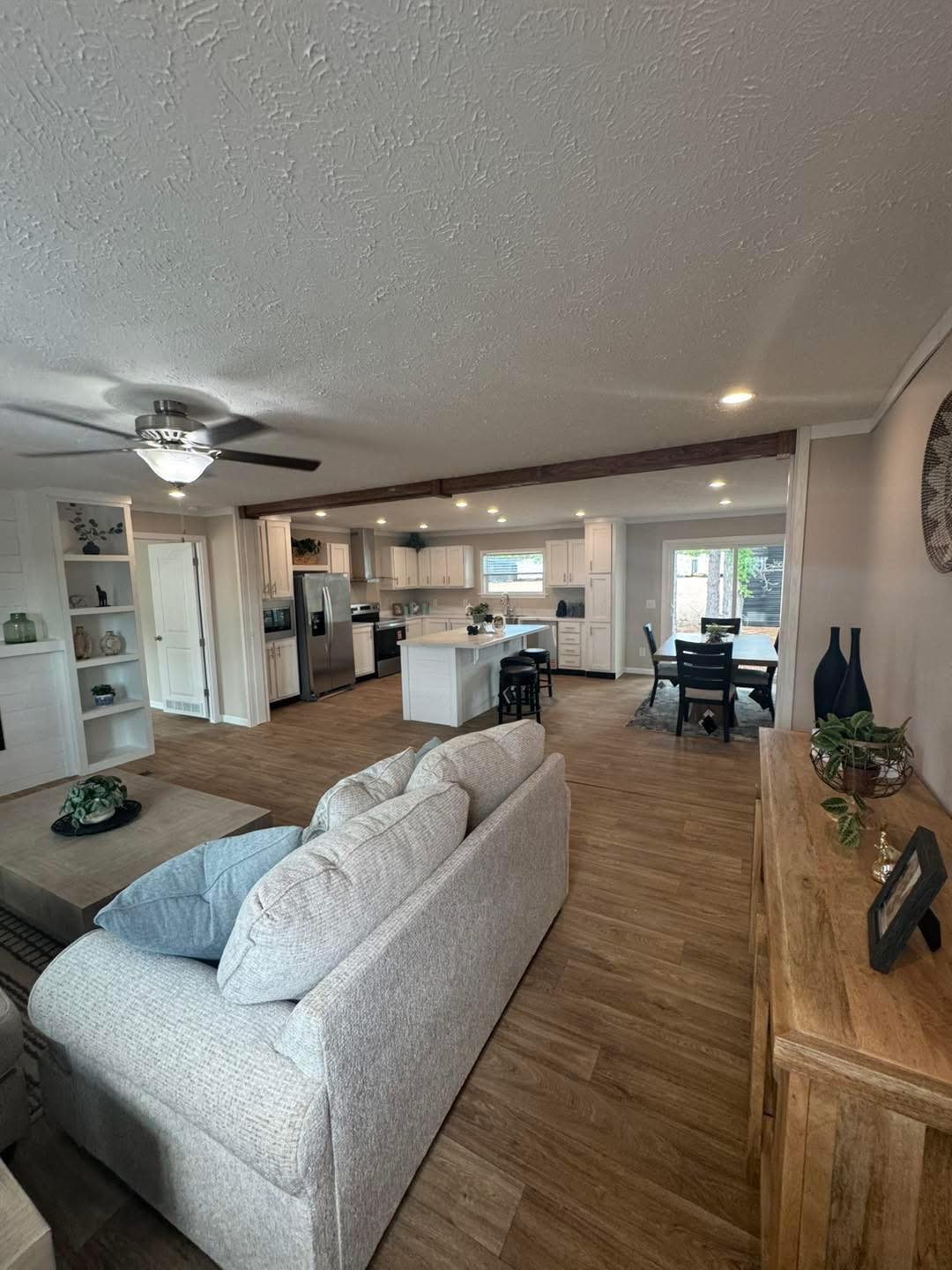 Spacious open-concept living area with modern kitchen, large beige sofa, wood floors, ceiling fan, and dining table. Bright and inviting atmosphere.