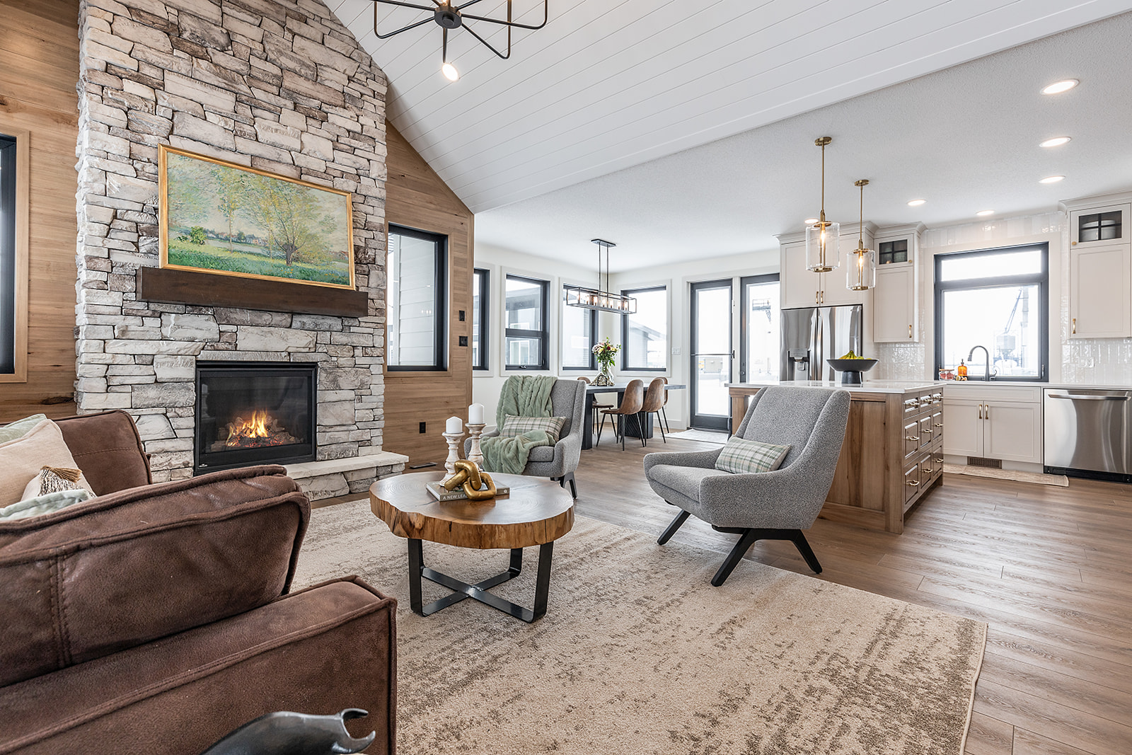 A modern living room with a stone fireplace, cozy armchairs, and a wooden coffee table. Open concept kitchen and dining area, creating a warm, inviting space.