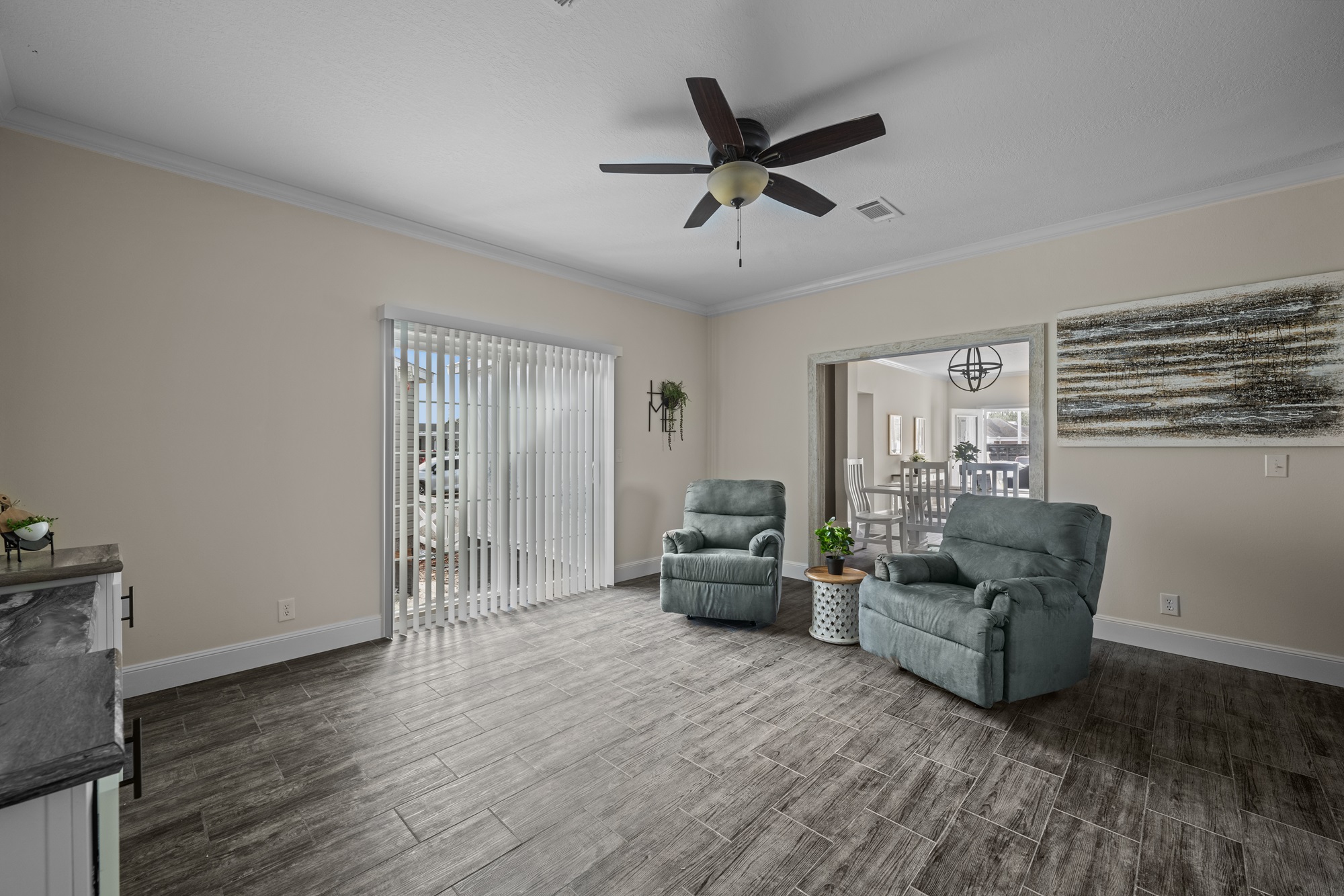 Living room with two teal recliners on wood-style flooring, a ceiling fan above. Bright sliding glass door and abstract art on a beige wall. Peaceful ambiance.