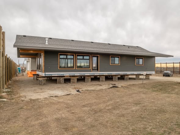 Elevated gray modular house on wooden blocks in a barren landscape, with cloudy skies; evokes a sense of transition and construction progress.