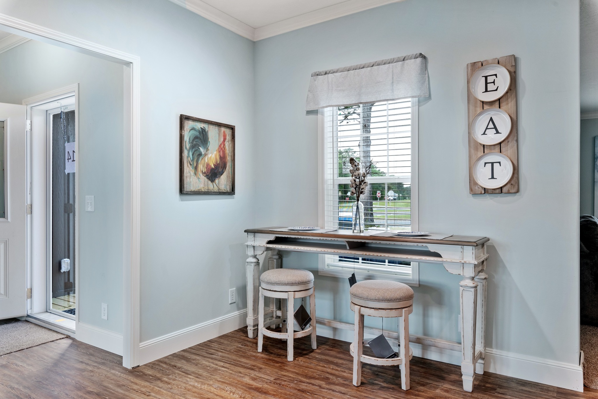 Cozy corner with light blue walls, featuring a rustic table, two cushioned stools, rooster painting, and "EAT" sign on wood. Bright and inviting atmosphere.