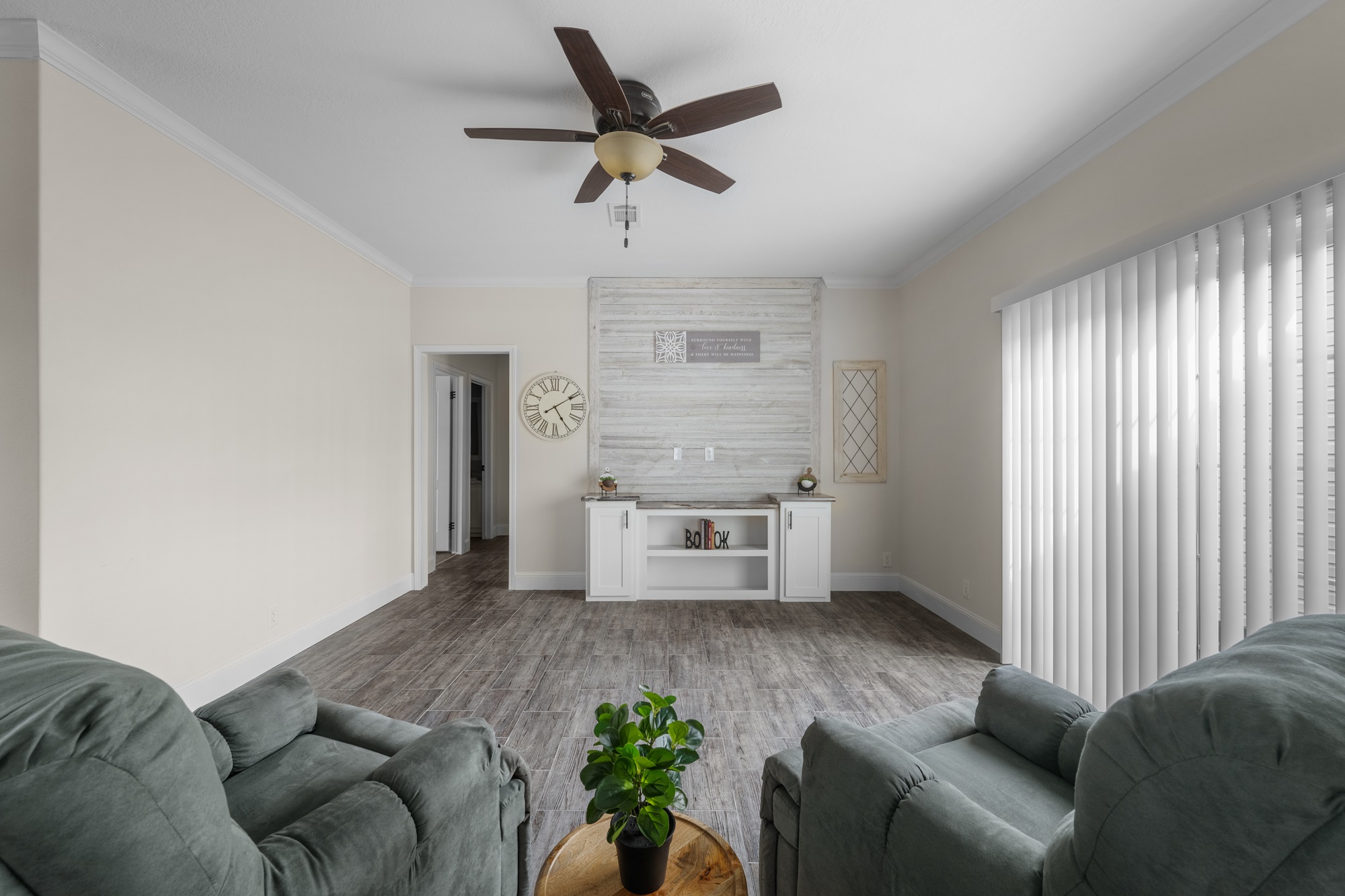 Bright living room with two green recliners facing a wooden accent wall, beige walls, a ceiling fan, and wide blinds. A plant on a round table adds warmth.