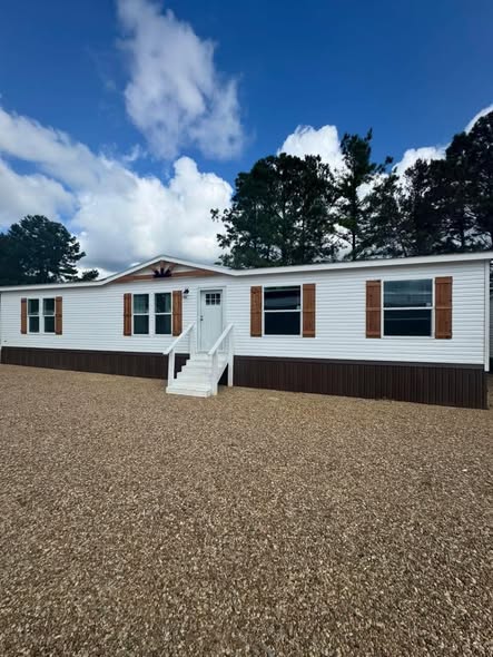 A white modular home with brown accents, sitting on a gravel lot under a partly cloudy blue sky. Surrounded by tall pine trees, it exudes tranquility.