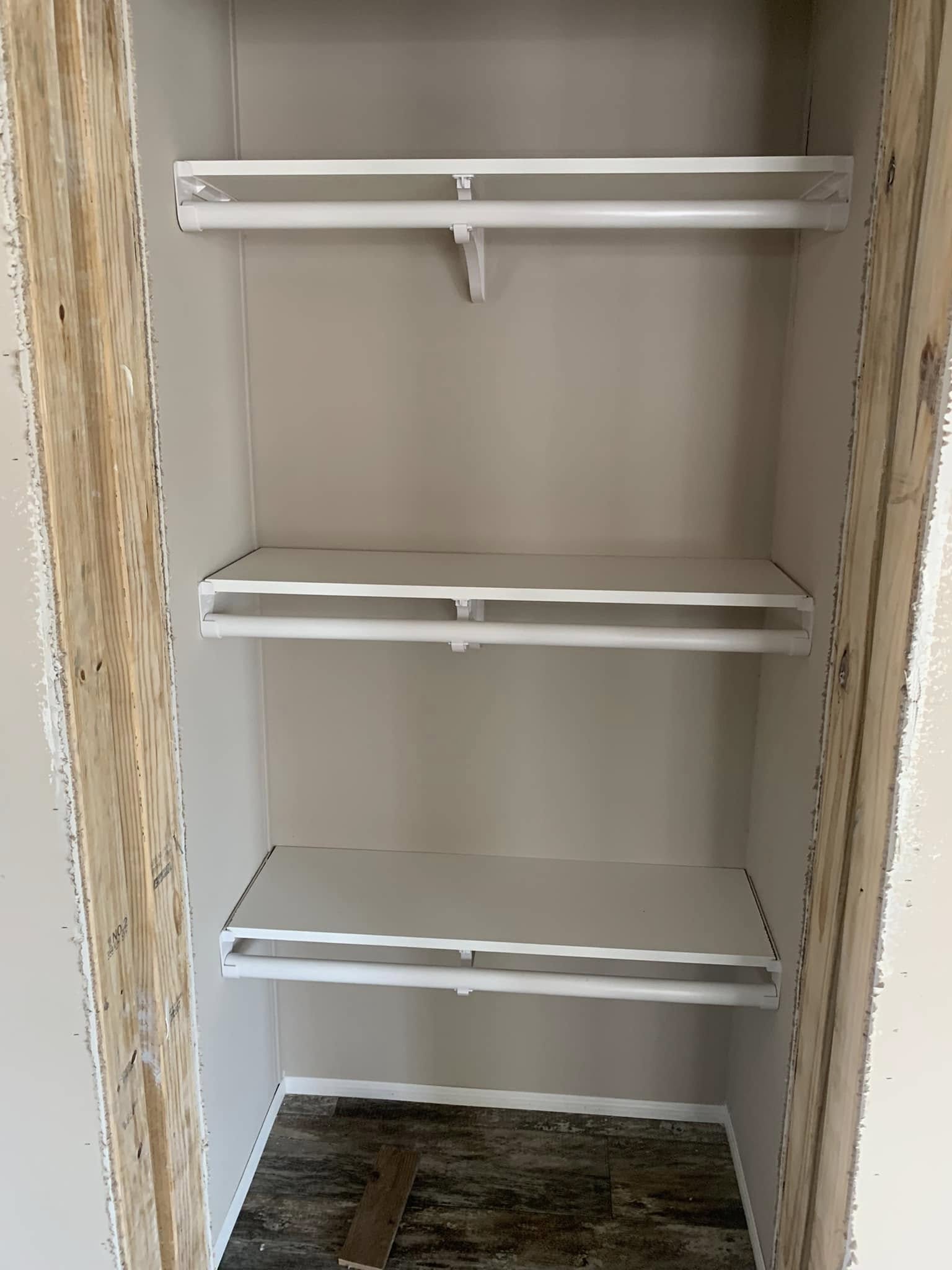 An unfinished closet with three white shelves and a wooden frame. The floor is wooden, and the walls are unpainted, conveying an incomplete look.