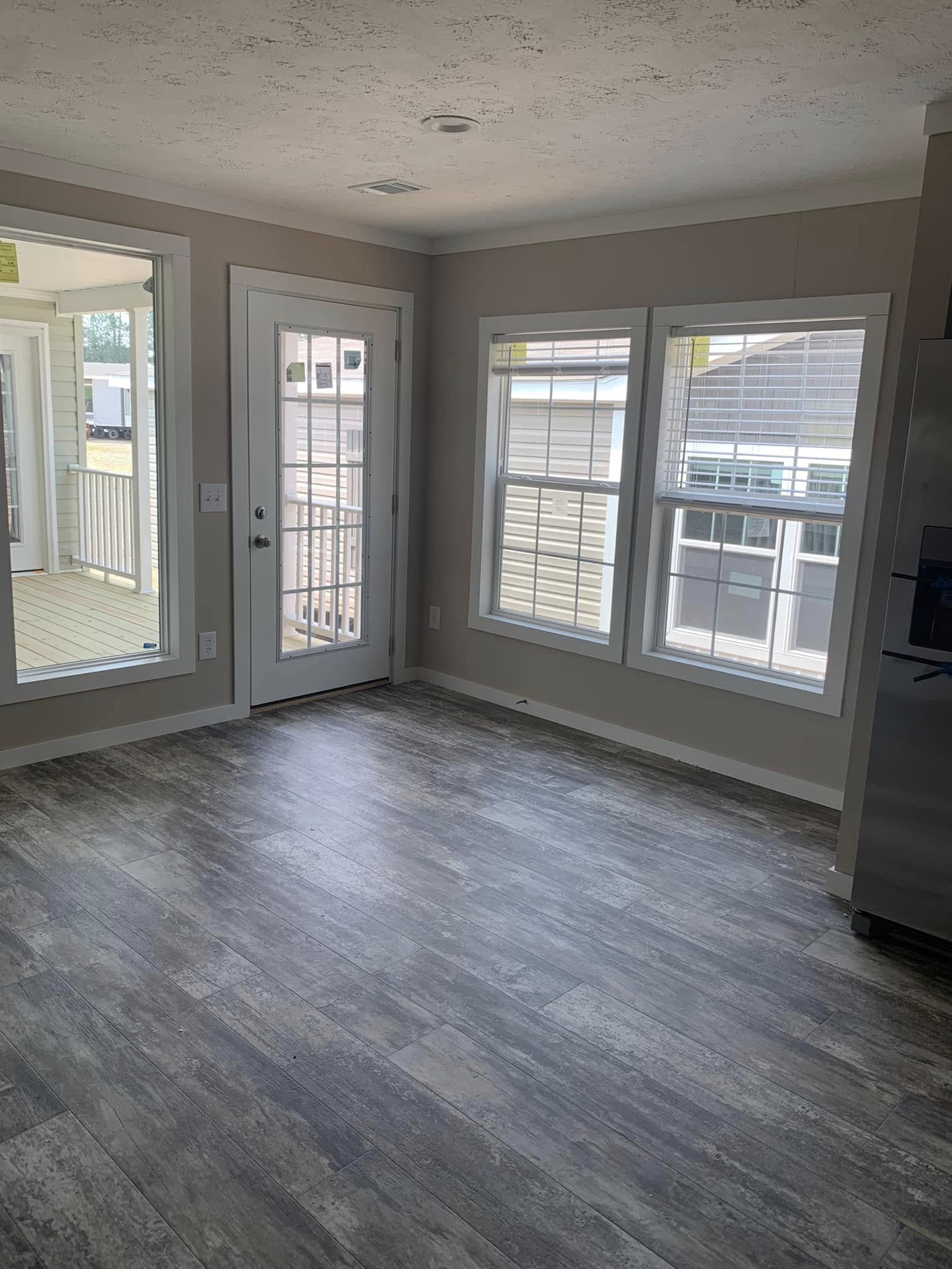A bright room with large windows and a glass door leading to a porch. The space has light gray wooden flooring and neutral walls, creating an airy atmosphere.