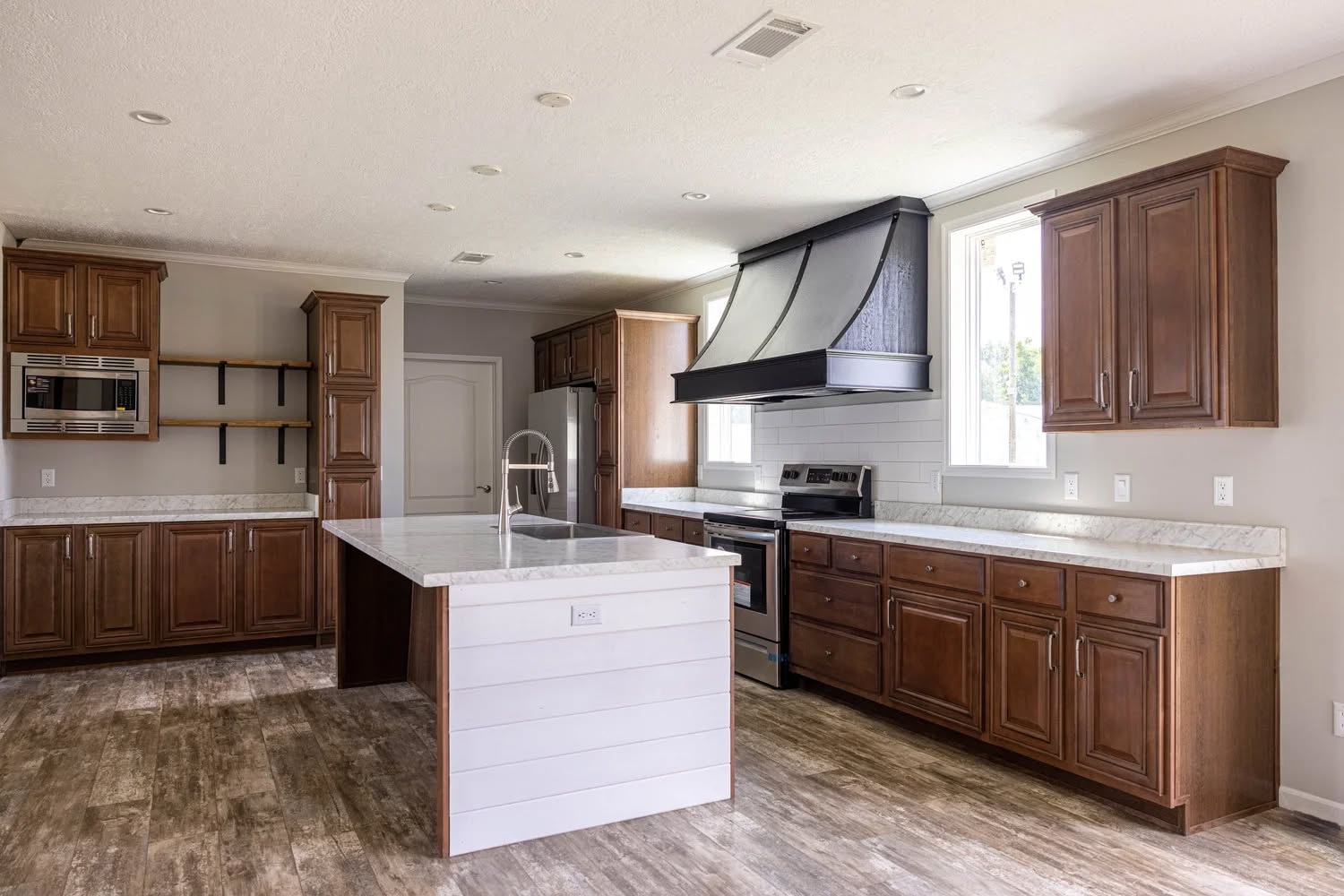 Spacious kitchen with rustic wood cabinets, white marble countertops, and a large island with a modern faucet. Light wood flooring and bright window.