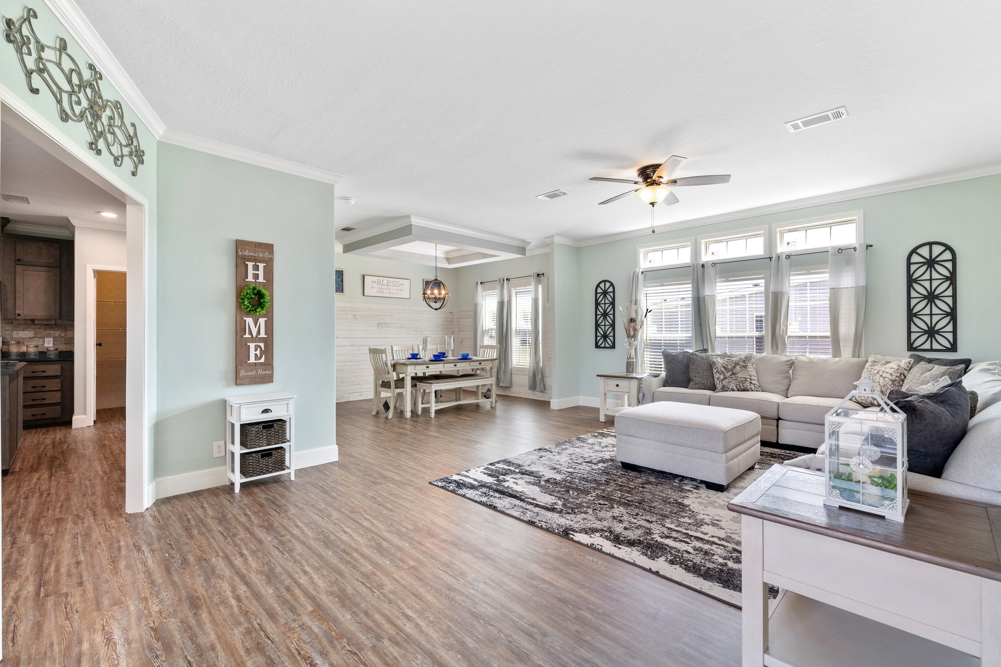 Spacious living room with light green walls, large gray sectional sofa, and decorative elements. Adjacent dining area with white table. Cozy, inviting ambiance.