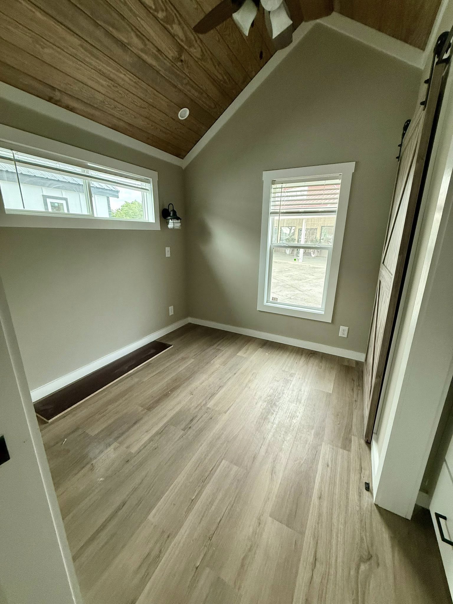 Small room with wooden ceiling and floor, beige walls, and two windows. A ceiling fan hangs above. The space feels cozy and bright.
