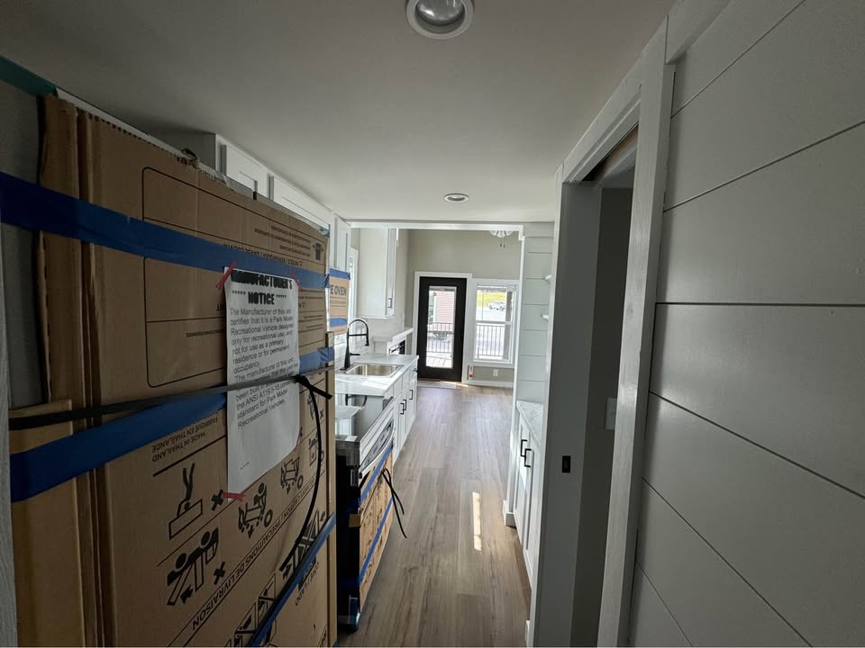Narrow kitchen with white cabinets, stainless steel sink, large appliance boxed, and natural light from a glass door. Wood flooring adds warmth.