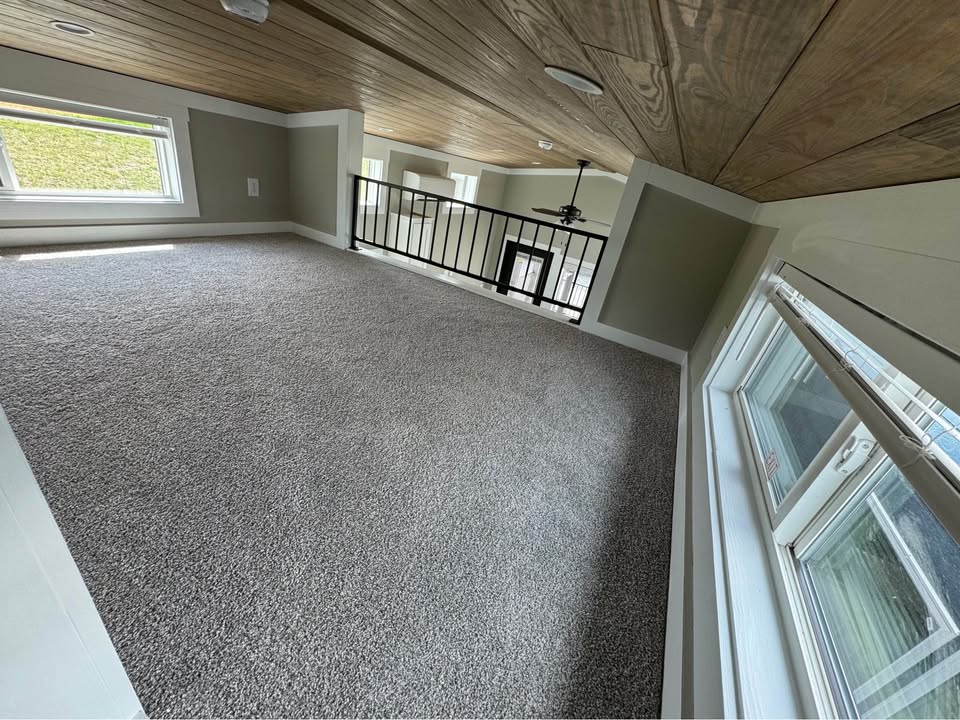Cozy loft space with gray carpet, wood ceiling, and windows on both sides. Overlooks room below with black railing, bright and inviting.