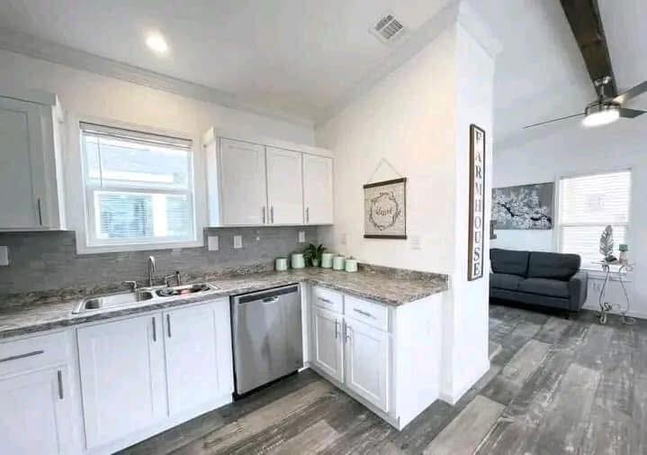 Bright kitchen with white cabinets and gray countertops has a farmhouse décor. The adjacent room has a dark sofa, modern art, and a ceiling fan.
