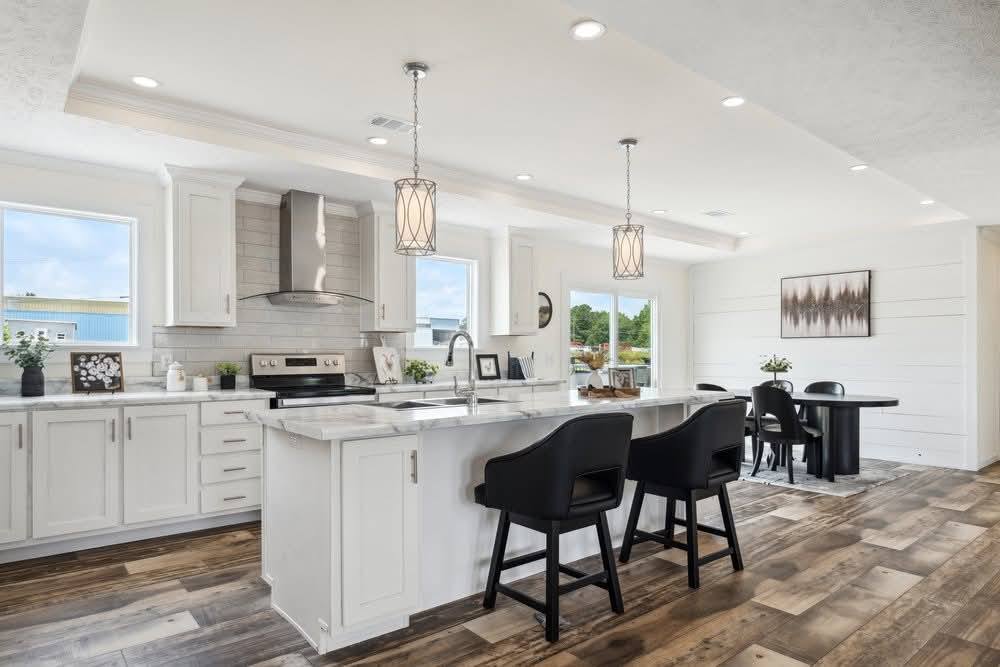 Modern kitchen with white cabinets, marble countertop, and hardwood floors. Black chairs at island, pendant lights, and adjacent dining area. Bright and airy.